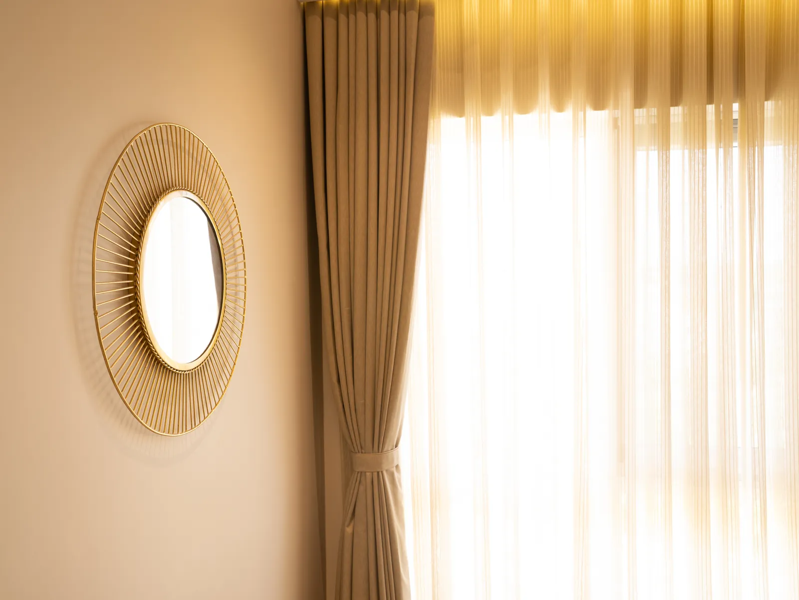 Round rattan mirror beside sheer curtains — Amlan's Residence